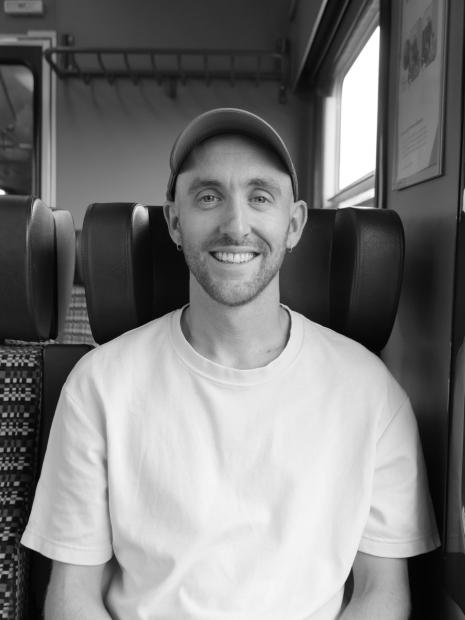 Black-and-white portrait of a smiling person sitting on a train. The person wears a light-colored cap, a light T-shirt, and small earrings, looking directly at the camera. In the background, there are padded train seats, a window with daylight, and a luggage rack.