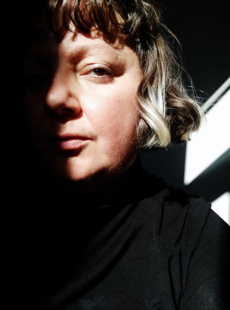Nahes Porträt einer Person mit kurzem, leicht gewelltem Haar und dunklem Oberteil. Ein starker Lichtstreifen fällt über eine Gesichtshälfte, während die andere Seite fast ganz im Schatten liegt. Im Hintergrund zeichnen sich helle, diagonale Lichtformen vor dunkler Fläche ab.