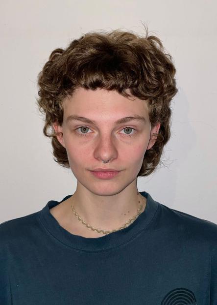 Portrait of a person with short, curly light-brown hair in a mullet style. The person looks straight into the camera, wearing a silver septum ring and a slim, braided-looking necklace. A loose, dark teal crew-neck top is visible. The background is a plain, light-coloured wall without additional details.