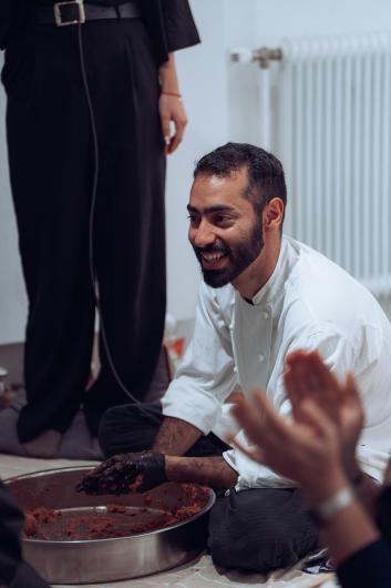 A bearded person kneels on the floor beside a large metal bowl filled with a reddish mixture. They wear a light-colored top and a black glove and are smiling. Blurred clapping hands are visible in the foreground, and another person stands in the background.