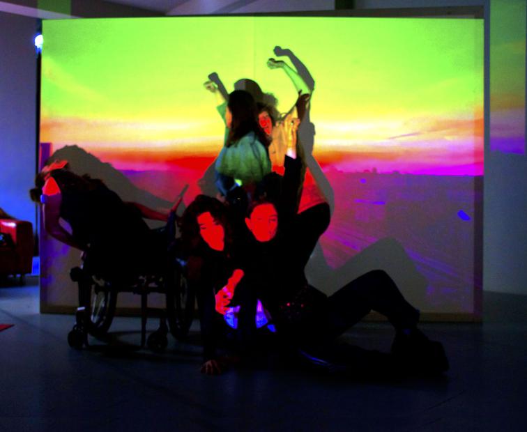 Eine größere Gruppe von Performer:innen posiert eng beieinander vor einer Wandprojektion in Neonfarben – grelles Grün, Gelb, Pink und Blau erinnern an eine Landschaft im Sonnenuntergang. Einige Körper sind deutlich zu sehen, andere verschwimmen in Bewegung, rechts vorne liegt eine Person am Boden, während sich im Hintergrund die Schatten von Körpern übereinanderlegen.