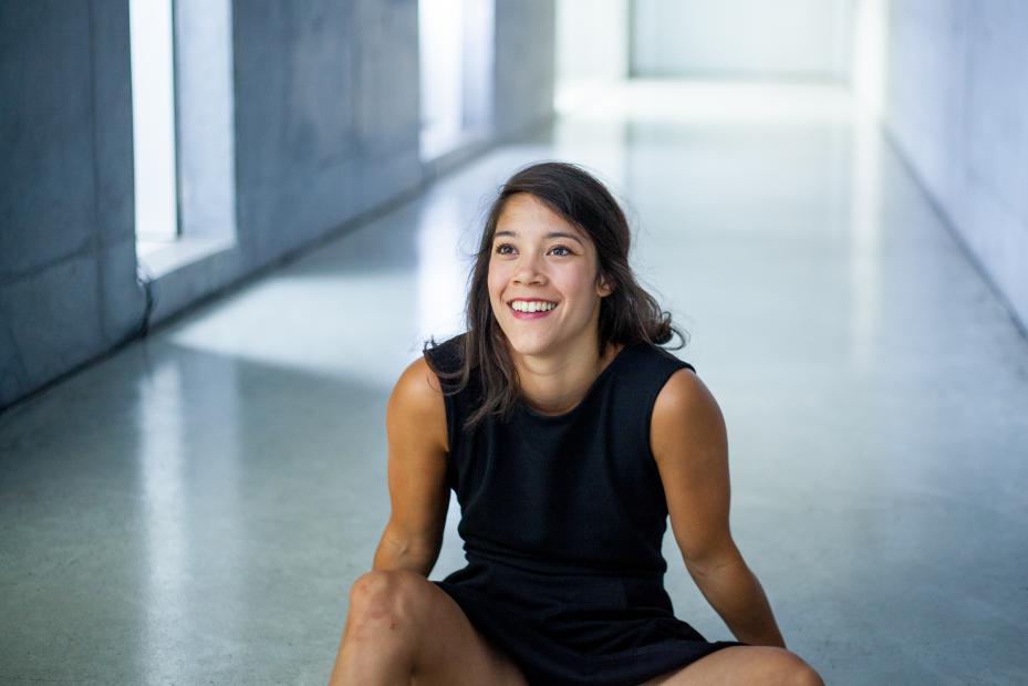 M. Pasman sitzt lachend auf dem Boden eines hellen Flurs. Die Person trägt ein ärmelloses schwarzes Kleid, sitzt locker mit angewinkelten Beinen, umgeben von glatten Wänden und Licht von den Fenstern im Hintergrund.