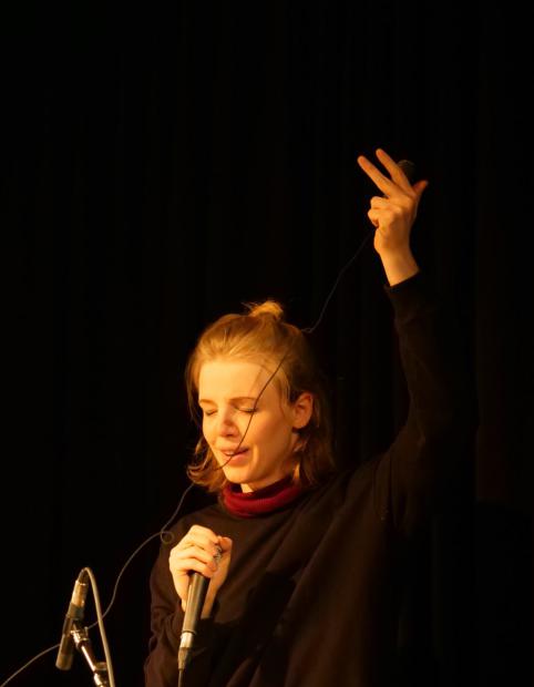 A person stands against a black background in warm stage light, eyes closed, as if singing or listening with deep concentration. They hold a microphone in one hand near the body, while the other arm is stretched high above the head. The microphone cable runs diagonally across the face and up towards the raised hand. The person is wearing a dark top with a high burgundy collar. The scene feels calm, intimate and charged with musical focus.