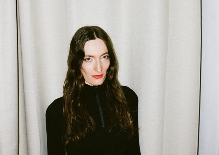 A person with long, wavy brown hair wears a black, form-fitting top with a zipper at the collar. She has a serious yet slightly mysterious facial expression with red lips. The background consists of a beige curtain.