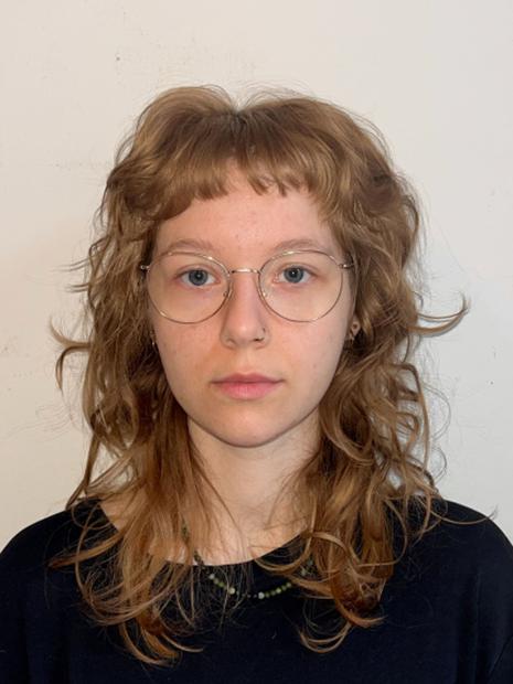 Portrait einer Person mit schulterlangem, welligem hellbraunem Haar und kurzem Pony. Die Person trägt eine runde, dünn gerahmte Brille, einen kleinen Nasenring und Ohrringe. Ein dunkles Oberteil mit Rundhalsausschnitt und eine feine Perlenkette sind zu sehen. Der Hintergrund ist eine einfarbige, helle Wand.
