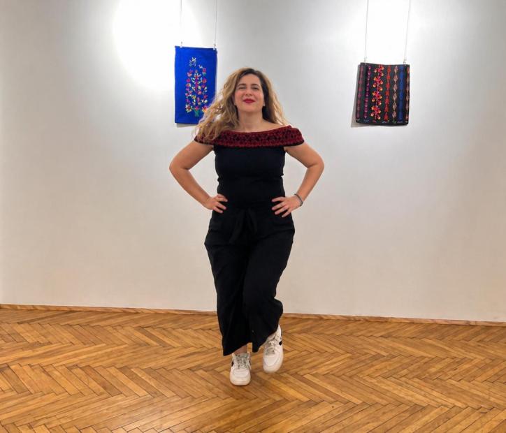 Eine Person steht mittig in einem hellen Ausstellungsraum auf einem Fischgrätparkett und blickt lächelnd in die Kamera. Sie trägt einen schwarzen Jumpsuit mit rotem, gemustertem Ausschnitt und weisse Turnschuhe, die Hände in die Hüften gestemmt. An der weissen Wand hinter ihr hängen zwei textile Bilder, eines blau mit floraler Stickerei, eines dunkel mit roten und blauen Ornamenten.