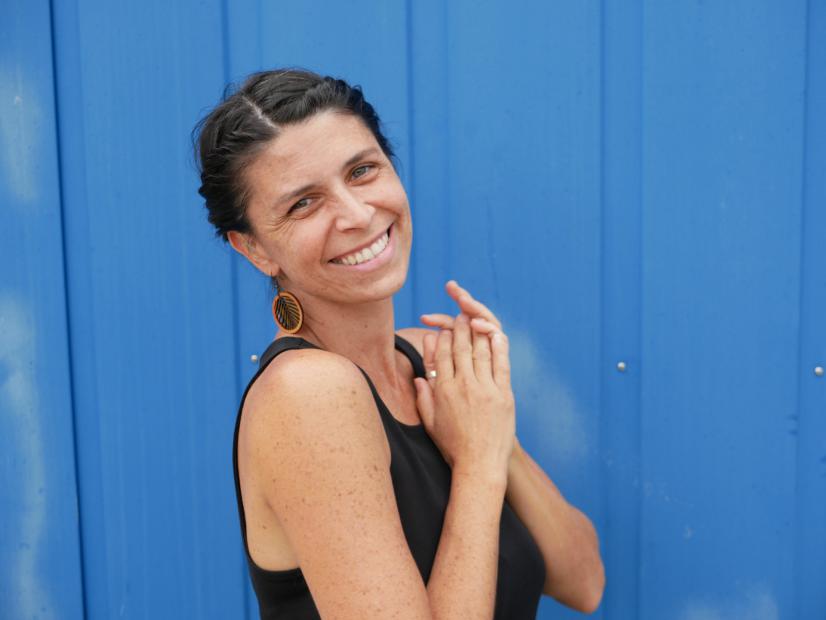 A person stands in front of a bright blue corrugated wall, smiling directly at the camera. Their dark hair is pinned back, and a round earring is visible on one side. They are wearing a black sleeveless top and holding their hands loosely in front of their chest, which gives the pose a soft, friendly feel. The daylight clearly highlights the face, the skin and the vivid blue background. The mood feels open, warm and welcoming.