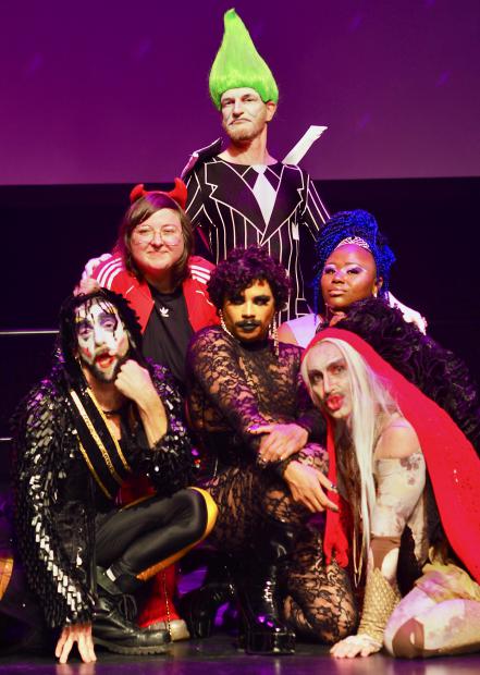 Gruppenporträt von sechs Personen auf einer Bühne, inszeniert wie ein Show-Finale. Die Personen tragen auffällige Kostüme, Make-up und Perücken in kräftigen Farben, darunter Glitzer, Spitze und ein leuchtend grüner, hochgestylter Haarschopf. Einige Personen knien oder sitzen im Vordergrund, andere stehen dahinter. Die Posen sind selbstbewusst, spielerisch und expressiv. Violettes Bühnenlicht verstärkt die theatralische, queere Ästhetik der Szene.