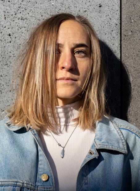 Porträt einer Person mit schulterlangem blondem Haar vor einer grauen Betonwand. Die Person trägt eine helle Jacke aus Jeansstoff über einem weißen Rollkragenoberteil und eine feine Halskette. Sonnenlicht fällt von der Seite auf das Gesicht und erzeugt starke Licht- und Schattenkontraste.