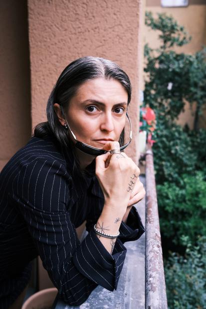 Portrait of a person on a balcony or exterior corridor with a blurred green courtyard behind them. They lean on a railing, look straight into the camera and hold a pair of sunglasses to their lips. They wear a dark pinstriped jacket, several rings and bracelets, and small tattoos are visible on the hand and forearm.