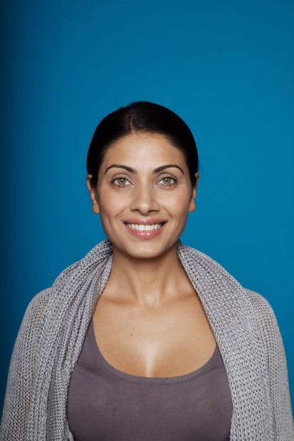 Portrait photo of a smiling person with dark hair pulled back and light-colored eyes in front of a plain blue background. The person is wearing a gray knitted shawl over a mauve top and is looking directly into the camera.