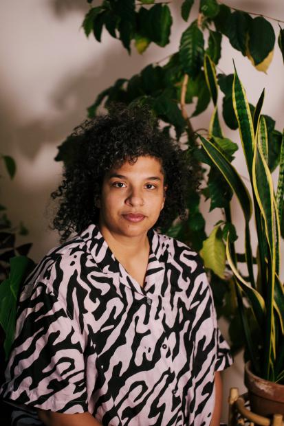 A person sits or stands calmly in front of several large houseplants, looking directly into the camera. They have dark curly hair and are wearing a loose-fitting shirt with a bold black and pink pattern. On the right side of the image, the tall upright leaves of a snake plant frame the scene, while more large green leaves fill the background. Warm light highlights the person’s face and patterned shirt, while the background remains soft and slightly shadowed. The mood feels calm, open and attentive.