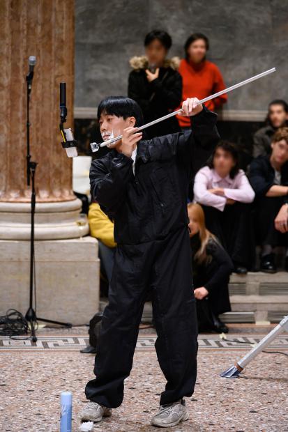 In einer großen Halle steht eine Person in einem schwarzen Overall und Turnschuhen auf einem dekorativen Steinboden. Die Person hat Rasierschaum um den Mund und hält einen langen Metallstab, an dessen Ende ein Rasierer befestigt ist, vor das Gesicht. Im Hintergrund sitzen mehrere Menschen auf Stufen und schauen zu, auf dem Boden stehen eine Spraydose, ein zerknülltes Papiertuch und ein Staubsaugerrohr, daneben befindet sich ein Mikrofonständer vor einer Marmorsäule.