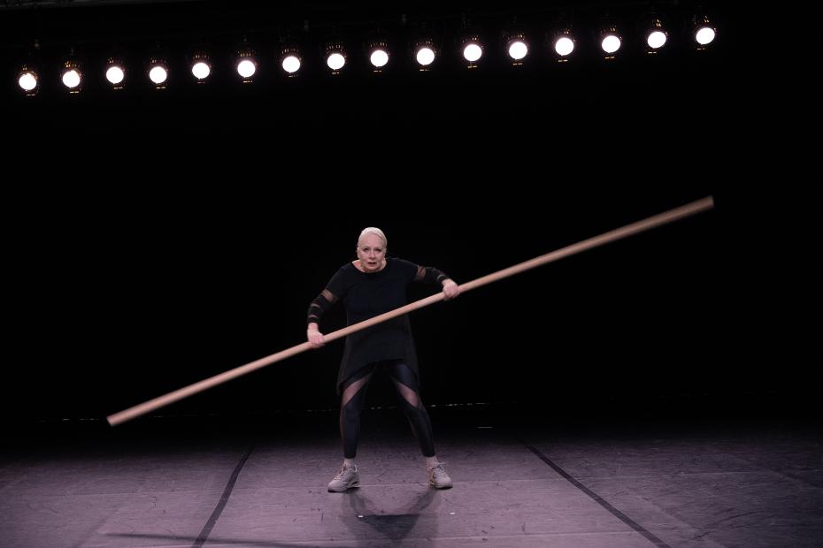 A person stands alone on a large, dark stage, holding a long pole diagonally across the space. Above them, a row of bright stage lights shines overhead. The person wears black performance clothing with sheer sections and light-coloured trainers. Their stance appears strong and focused.