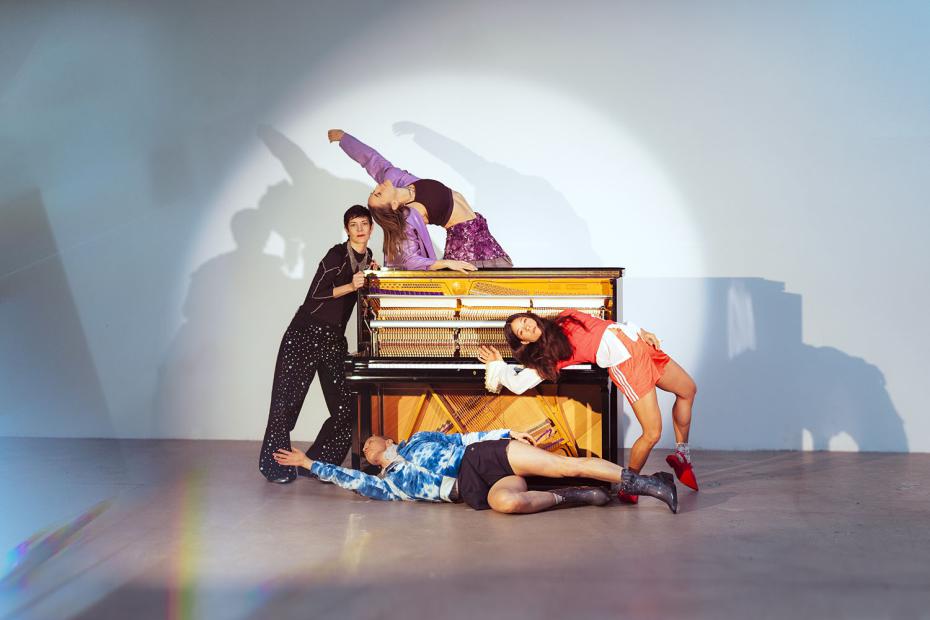 Vier Personen gruppieren sich dynamisch um ein geöffnetes Klavier im Studio. Eine Person in schwarzem Oberteil und gepunkteter Hose steht links und hält sich am Klavier fest. Oben auf dem Instrument beugt sich eine Person in kurzer, lilafarbener Jacke und gemustertem Rock weit nach hinten. Rechts lehnt eine Person in roter Sportkleidung seitlich an der Klavierfront, während eine Person in blauer Kapuzenjacke und kurzer Hose ausgestreckt am Boden vor dem Instrument liegt. Ein runder Lichtkegel hebt die Gruppe vor der hellen Wand hervor.