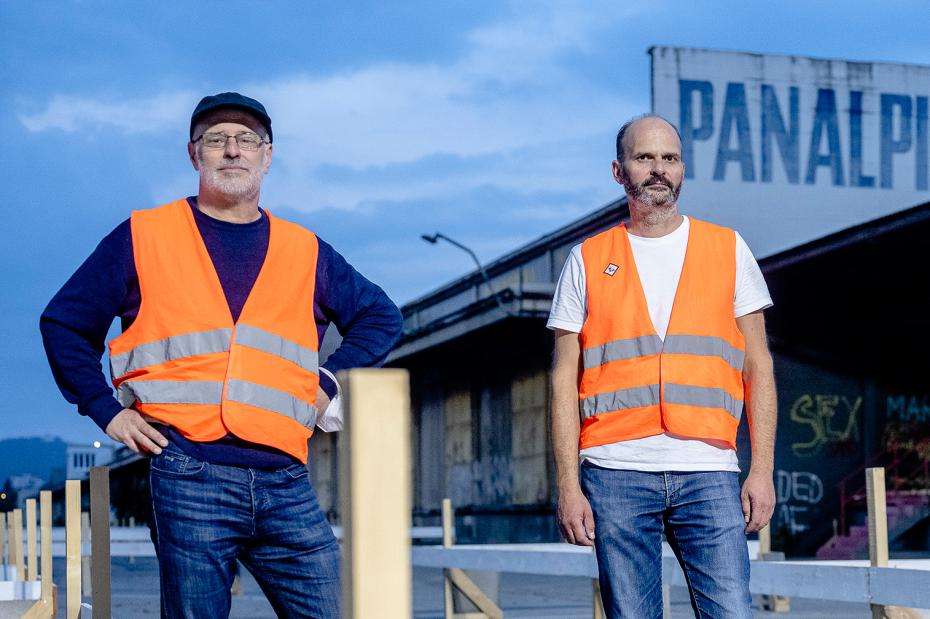 Zwei Personen stehen im Freien vor einem Industriegebäude in der Dämmerung und tragen orange Warnwesten über Alltagskleidung. Eine Person mit Kappe und Brille steht mit einer Hand in der Hüfte, die andere blickt direkt in die Kamera. Im Vordergrund sind helle Holzpfosten zu sehen, im Hintergrund eine wolkige blaue Abendstimmung.