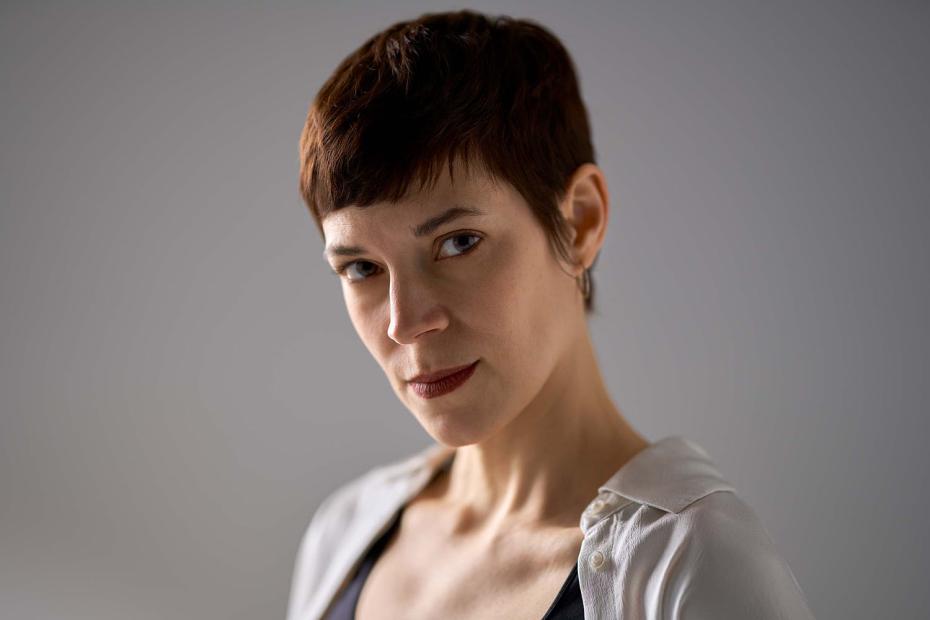 Close-up portrait of Verena Zeiner in front of a grey background. The person with short brown hair looks calmly into the camera, wearing a dark top with an open light shirt over it, soft light highlighting facial features and expression.
