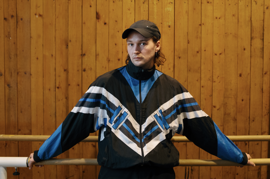 A person stands facing the camera in front of a wall made of vertical wooden panels, leaning casually with both arms on a ballet barre. The person wears a black cap and a sporty jacket in black, blue, and white. Their gaze is calm and direct, with a relaxed posture. The space resembles a dance studio or rehearsal room with warm, even lighting.