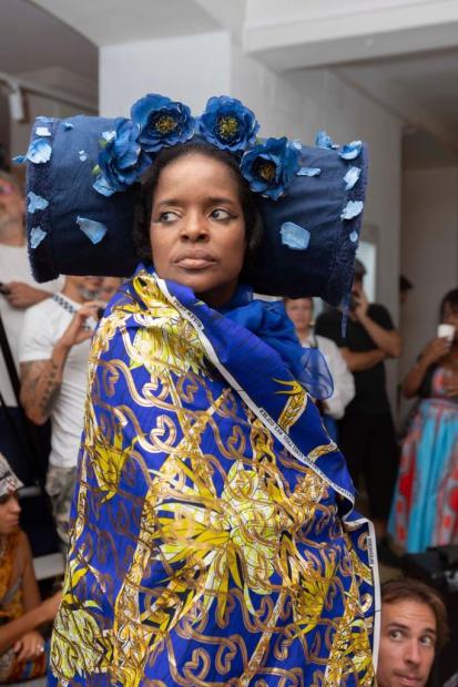 A person stands indoors, surrounded by an audience, looking seriously to one side. They are wearing a striking blue cylindrical headpiece decorated with several large blue flowers and scattered petals. Draped around their shoulders is a large blue, gold and white patterned fabric that almost completely wraps the body. The strong blue tones of the headpiece and garment stand out against the bright room and the softly blurred people in the background. The scene feels ceremonial, performative and intensely present.