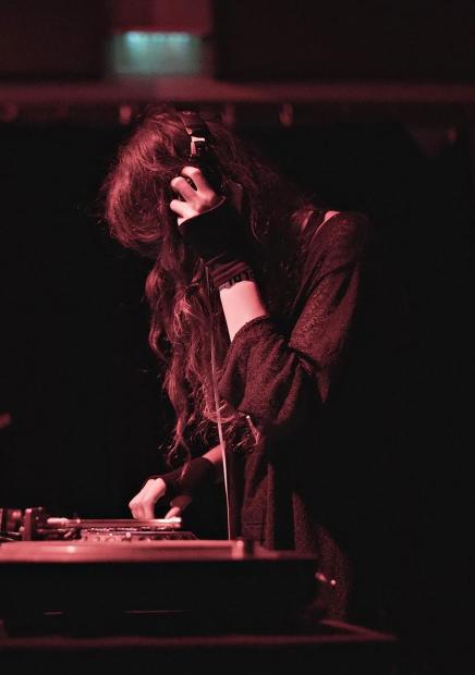 In einem dunklen Raum mit rötlich-pinkem Bühnenlicht steht eine DJ-Person hinter einem DJ-Pult. Die Person hat langes Haar, das teilweise ins Gesicht fällt, und trägt große Kopfhörer. Eine Hand hält die Kopfhörer an ein Ohr, während die andere Hand Bedienelemente am Mischpult berührt. Die Person trägt dunkle Kleidung mit langen Ärmeln. Das Gesicht ist nur teilweise sichtbar, da es von Haaren, Schatten und Licht verdeckt wird. Die Szene ist eng gerahmt, als würde man durch eine Öffnung oder zwischen Objekten hindurchblicken. Die Atmosphäre wirkt konzentriert, intim und clubtypisch, mit Fokus auf Soundarbeit, Körperhaltung und Präsenz.