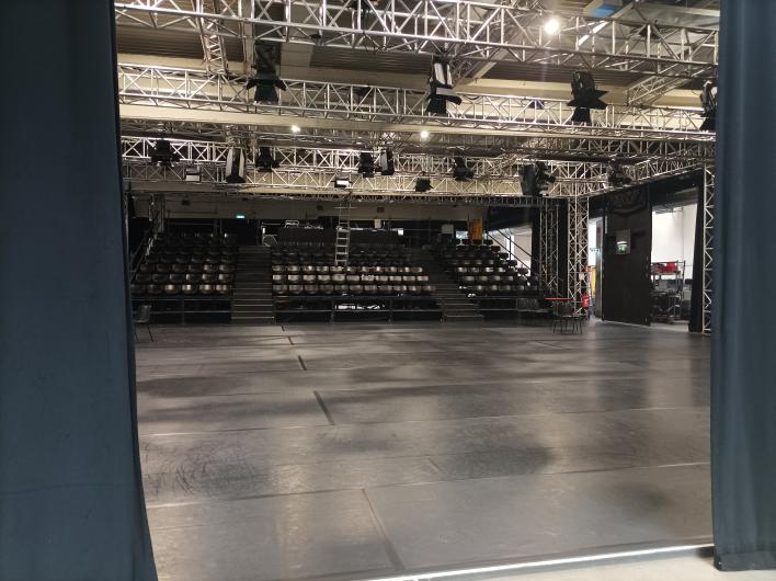 A view into an event hall with a black floor and a tiered audience stand with black seats. The hall is surrounded by a sturdy truss system, with numerous spotlights and speakers mounted on it. The space is framed by black curtains, which define the entrance area on the left and right. In the background, technical areas, chairs, and an emergency exit are visible. The atmosphere appears industrial and functional, ideal for theatre, concert, or event productions.