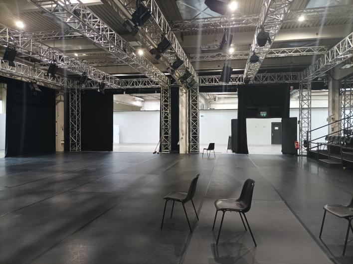 A large, black event space with a truss system on the ceiling, where numerous spotlights and speakers are mounted. The floor is covered with black mats, and several black plastic chairs are scattered throughout the room. Black curtains mark various entrances and transitions to adjacent areas. In the background, an open passage leads to a brighter, industrial-looking area with white walls and doors. On the right, there is a tiered seating area with black chairs. The atmosphere appears flexible and suitable for various event formats.