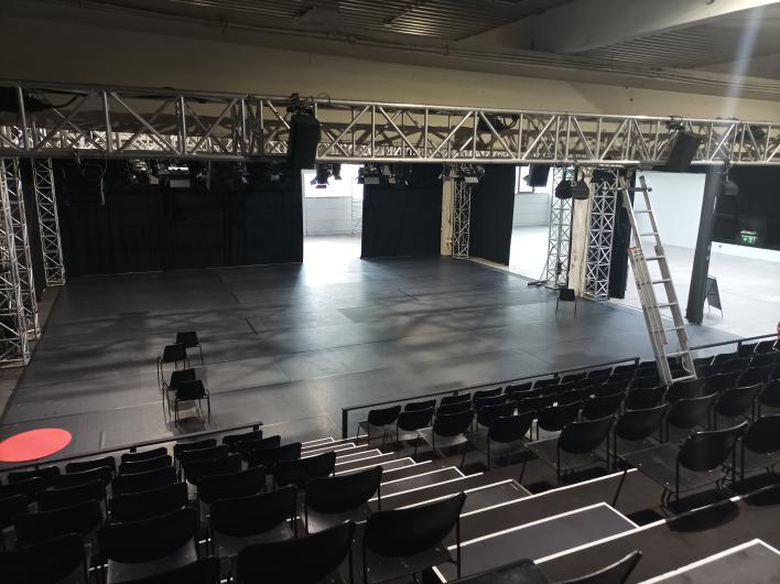 View from an elevated grandstand onto a large, black event space. The room is equipped with a ceiling truss system, with numerous spotlights and speakers attached. The floor is covered with black mats, and a few chairs are placed in the center. Black curtains mark various entrances. On the right side, a long ladder leans against the structure. In the background, open passages lead to a brighter, industrial area with white walls. The atmosphere is adaptable and suitable for various event formats.