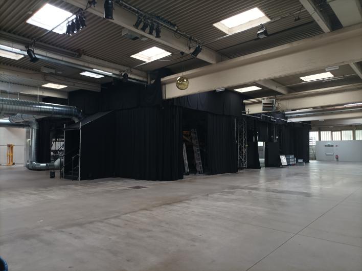 A large, industrial-looking space with a high ceiling, metal beams, and visible ventilation pipes. The walls are mostly in neutral colors. In the center, there is a modular stage construction draped in black, with black curtains, truss structures, and ladders. Several spotlights and a golden disco ball are mounted on the ceiling. The floor is made of smooth concrete, and natural light enters through skylights. In the background, additional technical and event equipment as well as an emergency exit can be seen.