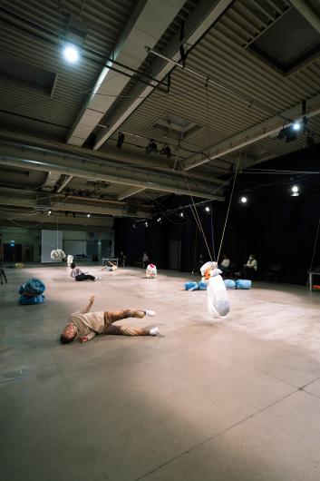 A large, open space with an industrial character, defined by a high ceiling with exposed metal beams, ventilation systems, and hanging lights. The floor is made of smooth concrete, and long black curtains hang on one side of the room. The lighting is functional, with multiple spotlights evenly illuminating the area. The space feels like a multi-purpose hall or an experimental venue for performance art.