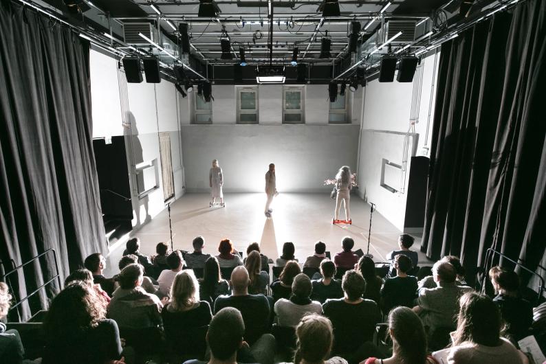 Der Raum ist ein kleiner, minimalistischer Theatersaal mit einer schlichten, modernen Ästhetik. Die Bühne hat eine helle, glatte Bodenfläche und wird von drei weißen Wänden umgeben, die keinerlei Dekoration aufweisen. Hinten an der Wand befinden sich drei hohe, schmale Fenster, die ein wenig natürliches Licht hineinlassen. Die Decke ist mit einem dichten Gitter aus Scheinwerfern, Lautsprechern und technischen Installationen versehen, die für die Beleuchtung und Akustik der Aufführung sorgen.  Der Zuschauerbereich besteht aus mehreren Reihen ansteigender Sitze, von denen aus das Publikum die Performance aus nächster Nähe beobachtet. An den Seiten hängen schwarze Vorhänge, die den Bühnenraum einrahmen und möglicherweise als flexible Bühnenelemente genutzt werden können.