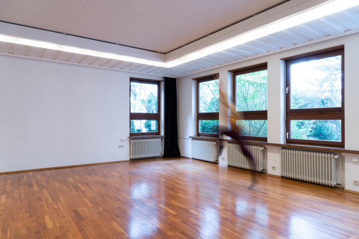 Ein heller, leerer Proberaum mit Holzparkett und großen Fenstern, durch die Tageslicht und grüne Bäume sichtbar sind. An der rechten Seite ist eine verschwommene Bewegung einer Person zu sehen, die sich schnell durch den Raum bewegt.